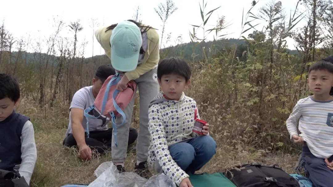 几个孩子和大人在长满高大杂草的荒地上,一名戴帽子的女性整理背包,几个孩子蹲坐或站立在周围。