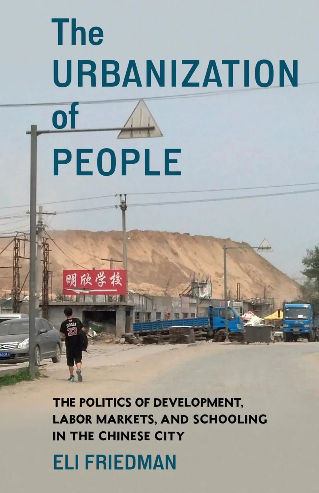封面照片为尘土飞扬的城郊道路,一名男孩背着书包独自走过,路边停着卡车,红色招牌写着“明欣学校”,背景是巨大的沙土堆;上方印着书名《The Urbanization of People》及作者 Eli Friedman。