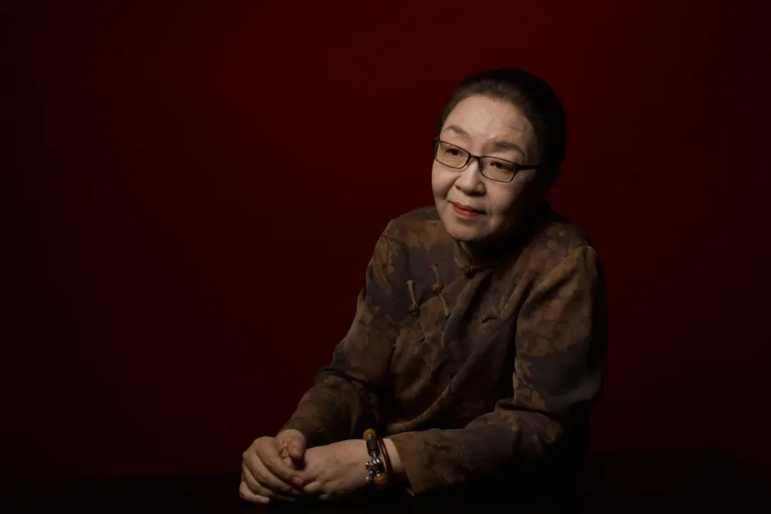 一位戴眼镜、穿深色暗纹上衣的女性坐姿肖像照,双手交叠置于桌面,背景为暗红色。
