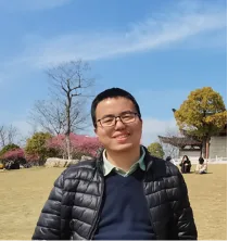 一名戴眼镜、穿黑色羽绒服的男子站在户外草地上留影,背景有樱花树与凉亭。