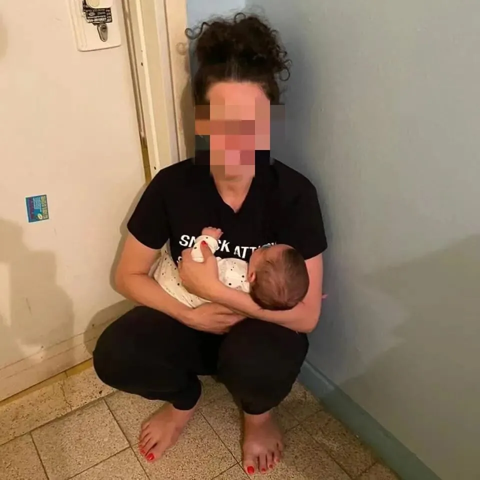 画面中女子面部被打码处理,赤脚蹲在房间墙角地上,双臂抱着一名婴儿,神情紧张地哄着孩子。