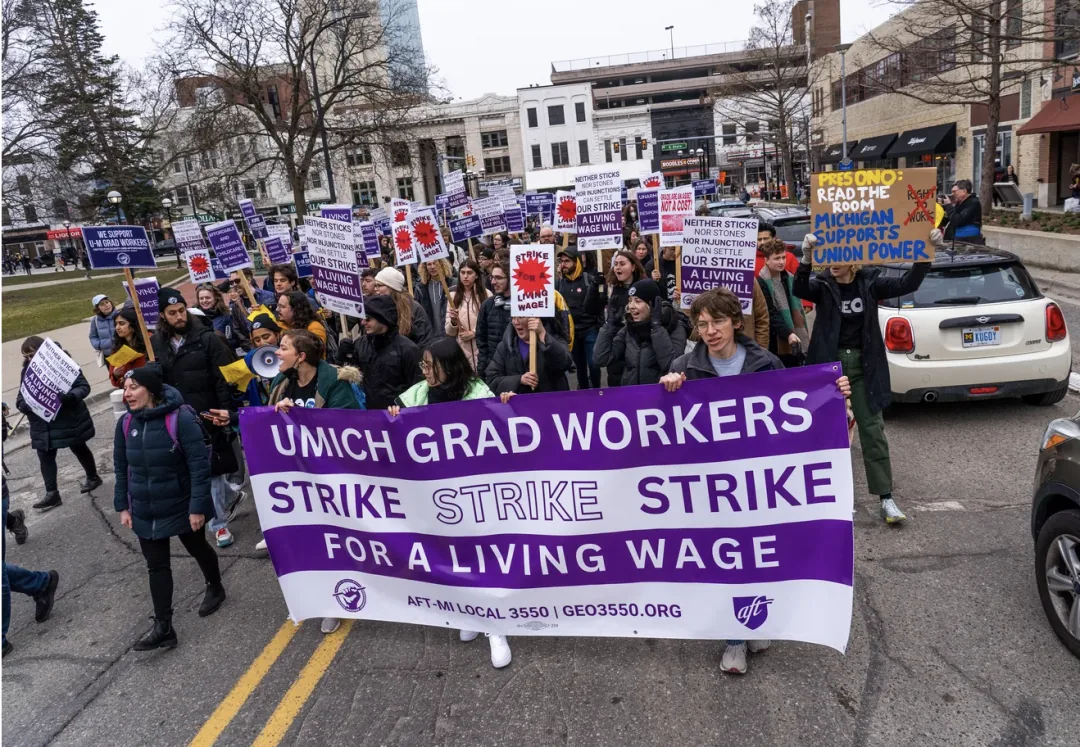 一大群研究生工人在街头游行,前排举着写有'UMICH GRAD WORKERS STRIKE STRIKE STRIKE FOR A LIVING WAGE'的紫白横幅,队伍中许多人举着标语牌,街道两旁是校园建筑。