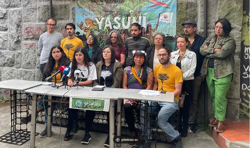 一群身穿黄色“Yasunidos”T恤的运动成员坐在摆有麦克风的桌前召开记者招待会,身后站着支持者,背景是画有雨林与动物的“Yasuní”横幅。