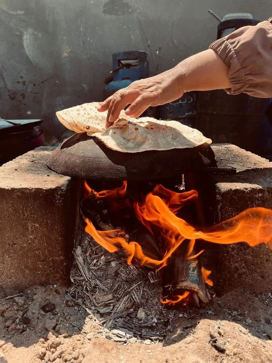 一只手正在砖砌炉灶上翻烤圆形面饼,炉灶下柴火燃烧着橙色火焰。