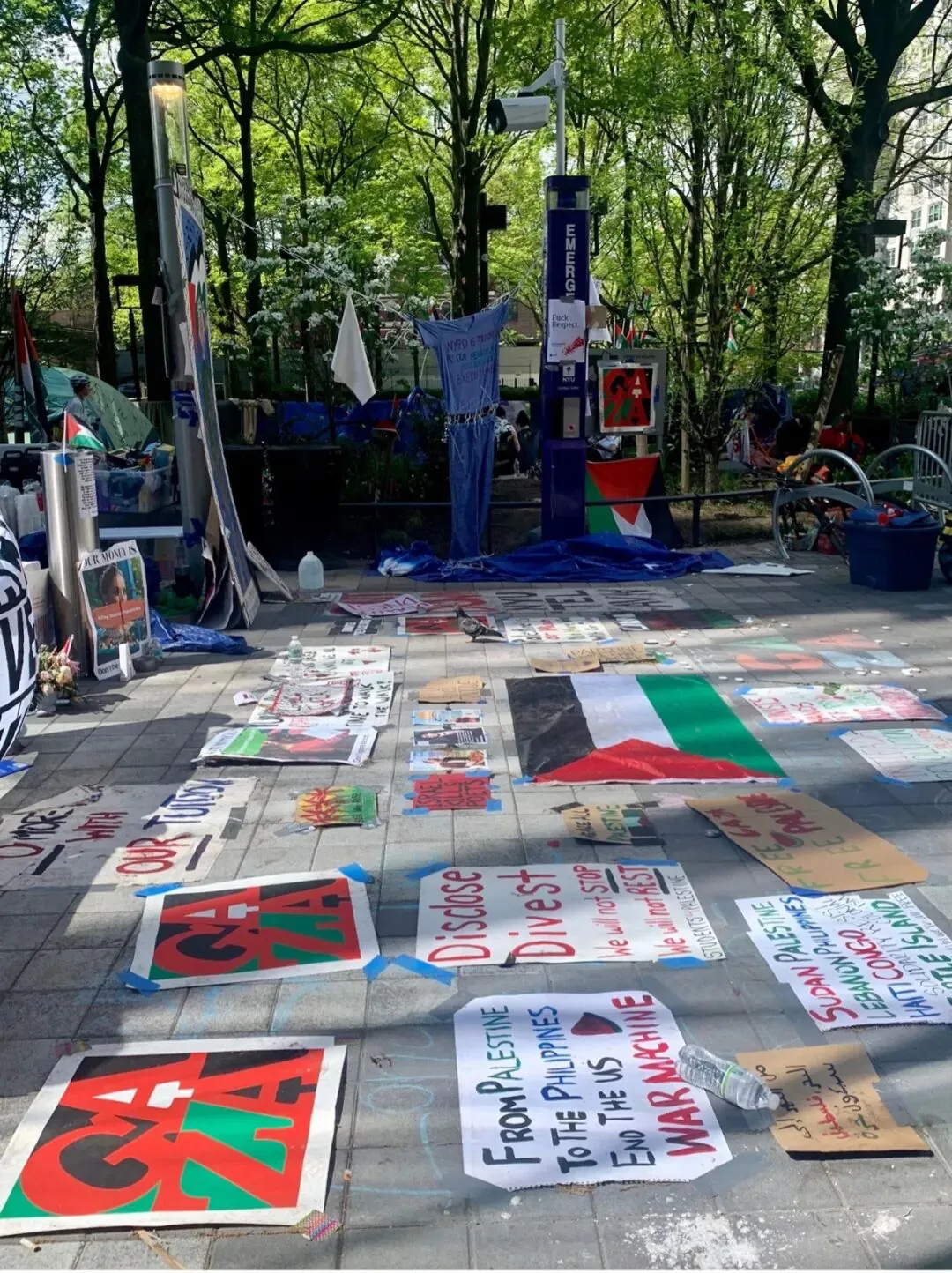 白天户外照片:广场地面上摆放着多张手绘标语牌,写着「GAZA」「Disclose Divest」「From Palestine to the Philippines, End the US War Machine」等,旁边有一面绘制的巴勒斯坦旗帜,周围是树木和帐篷区。