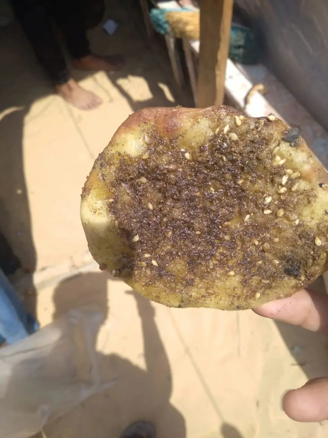 一只手举着一块表面撒满深色调料颗粒的扁面饼,背景为室外地面和一双光脚。