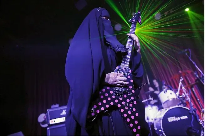 身穿黑色罩袍、蒙面的女性在舞台上弹奏粉色波点电吉他,背景有绿色激光灯与鼓手剪影。