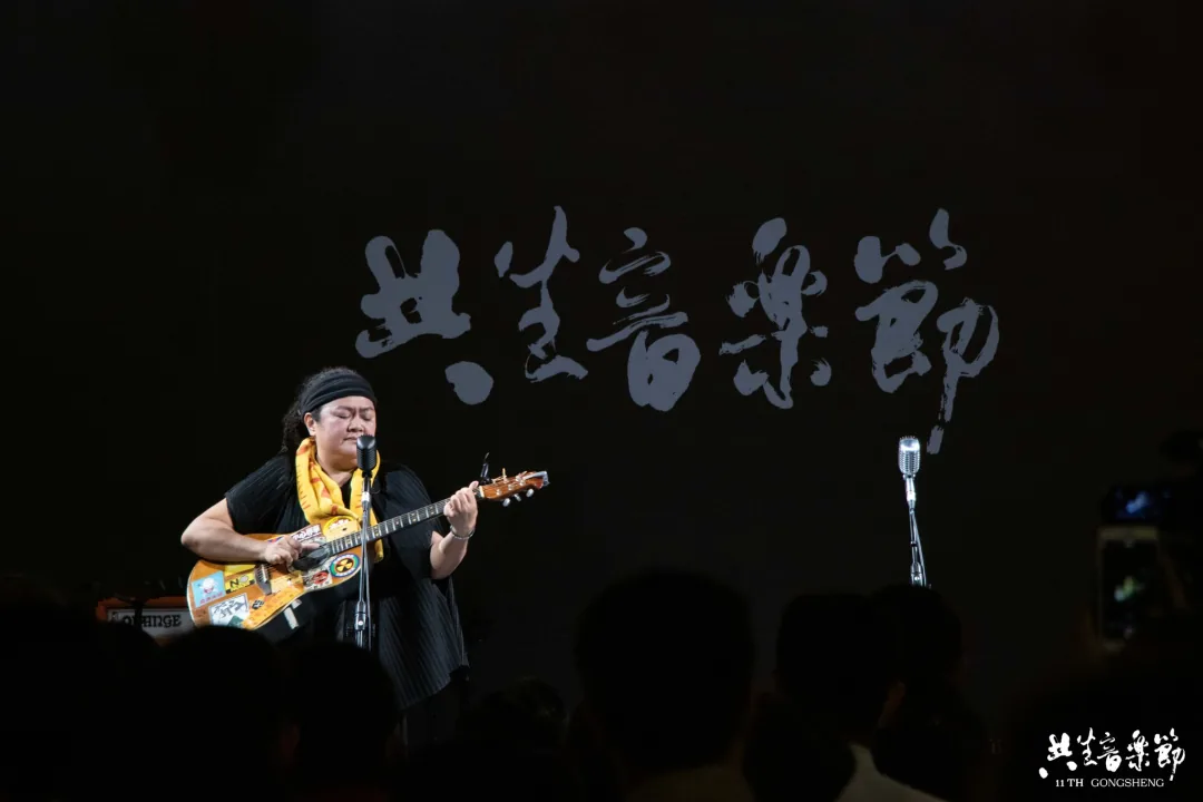 一位戴头巾、系彩色围巾的女性在台上抱着吉他弹唱,身后暗色背景写着“共生音乐节”字样。