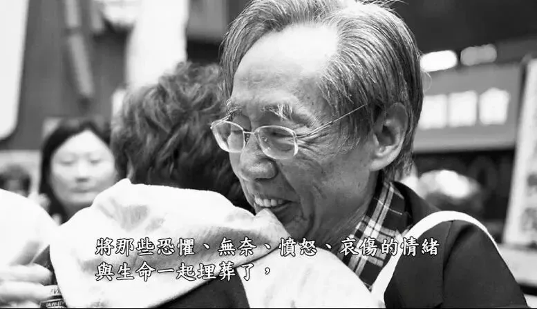 纪录片截图,黑白画面中戴眼镜的老年男性微笑着拥抱他人,画面字幕为“将那些恐惧、无奈、愤怒、哀伤的情绪与生命一起埋葬了,”