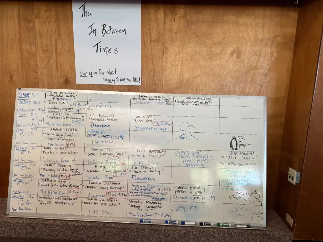 木墙上贴着白纸,手写标题“The In Between Times”,下书“Sign up in free slots! Show up to what you like!”,下方是一块贴满彩笔手写活动安排的日程白板,具体字迹密集潦草,多不可辨。