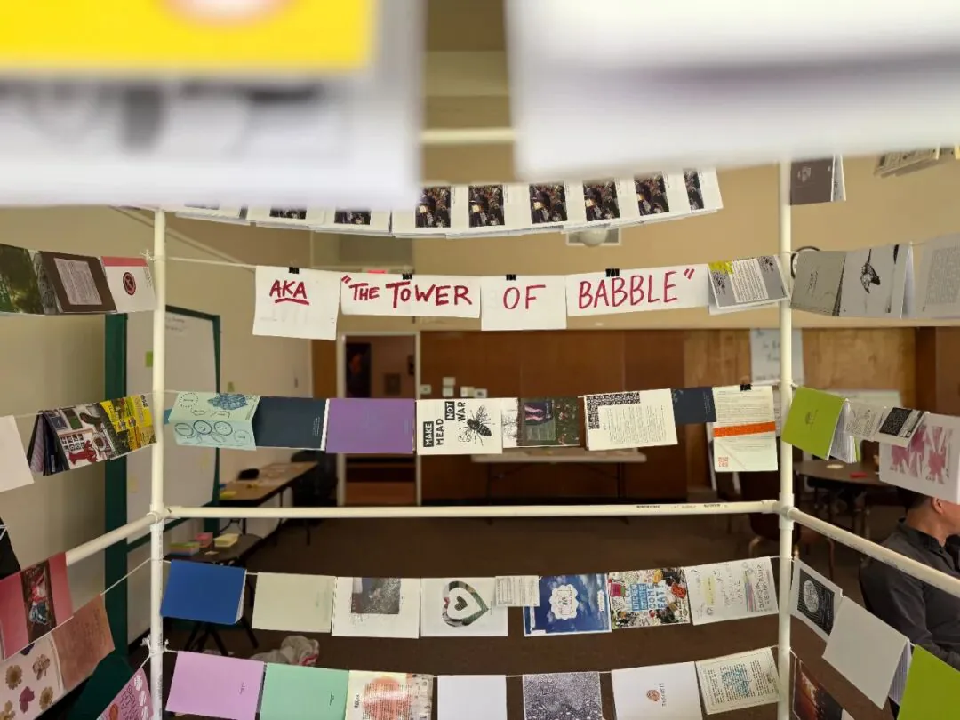 用PVC管搭成的展示架,多排彩色手工杂志用夹子悬挂展示,中间横幅手写“AKA 'THE TOWER OF BABBLE'”。