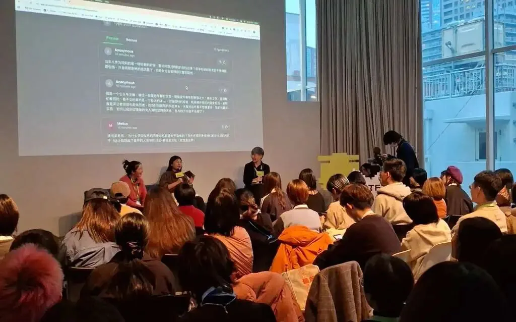 三位讲者站在台前对谈,身后大屏幕展示听众匿名提交的中文提问和留言,台下观众坐满整个空间,落地窗外可见城市高楼。