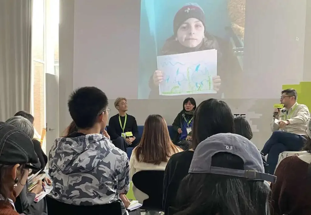 大屏幕播放一名戴毛线帽的孩子举着自己手绘图画的视频画面,台前几位讲者围坐对谈,台下观众围坐聆听。