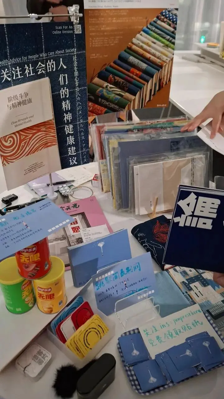 市集摊位桌面摆满自制小志、明信片和铁盒零食,海报副标题写着“阶级斗争与精神健康”(Class Struggle and Mental Health),旁桌还有蓝色手写卡片。