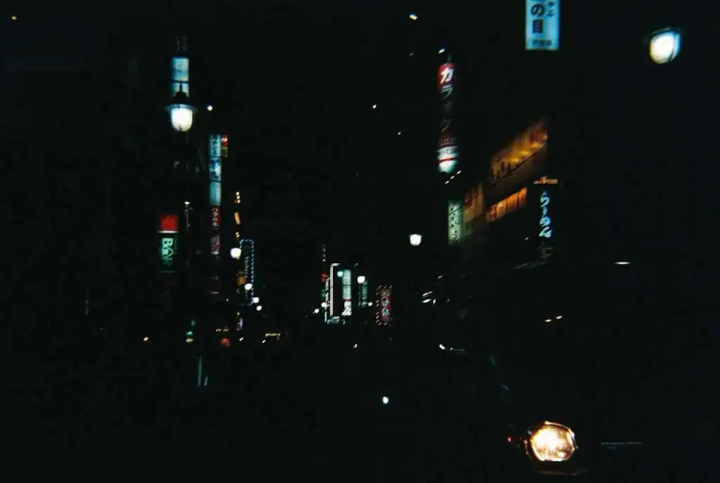 夜晚的东京街道,两侧竖立着多块发光的日文招牌,昏暗中一辆汽车的车头灯在画面右下角亮起。