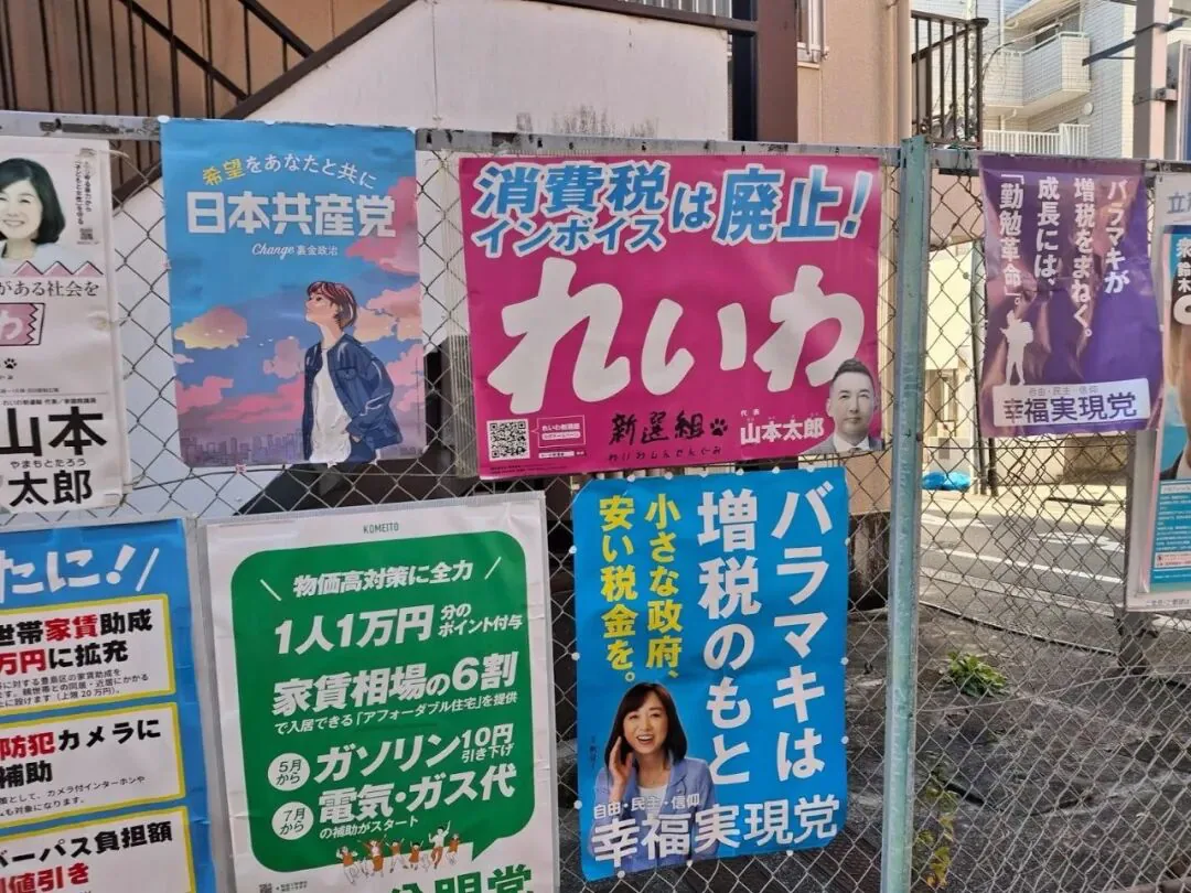 一面铁丝网围栏上贴满日本政党竞选海报,可见“日本共产党”、“れいわ”(配山本太郎头像,标语“消费税・発票废止!”)、“幸福実現党”等,背景是居民楼。