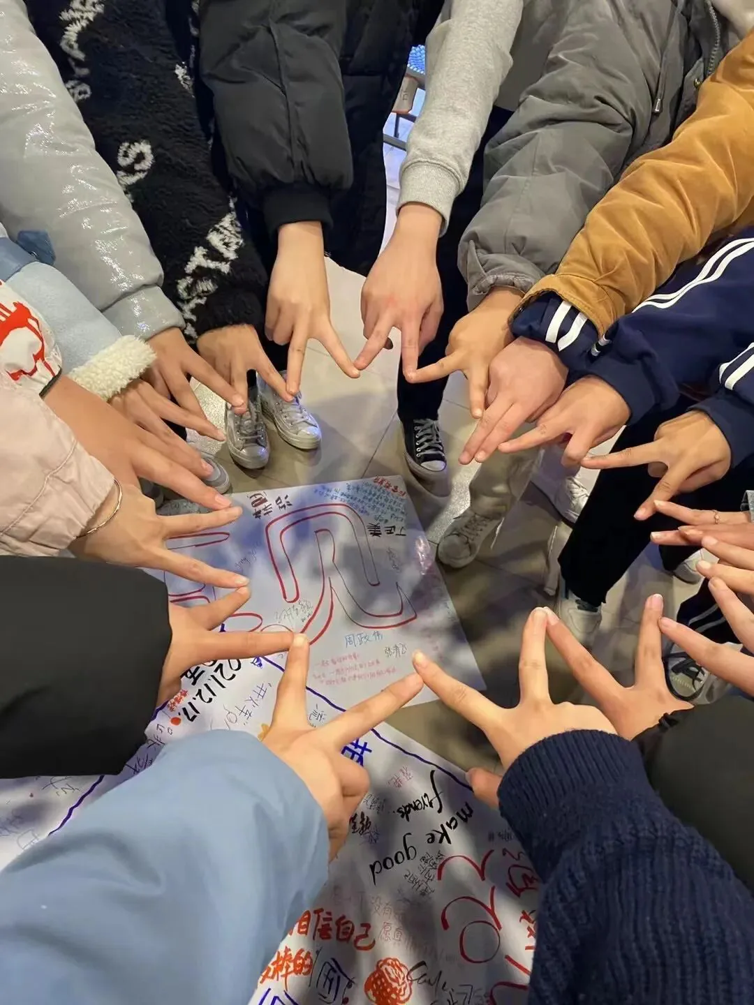 俯拍多人围成一圈,众多手指伸向中央写满彩色文字与签名的海报,做出团队仪式动作。