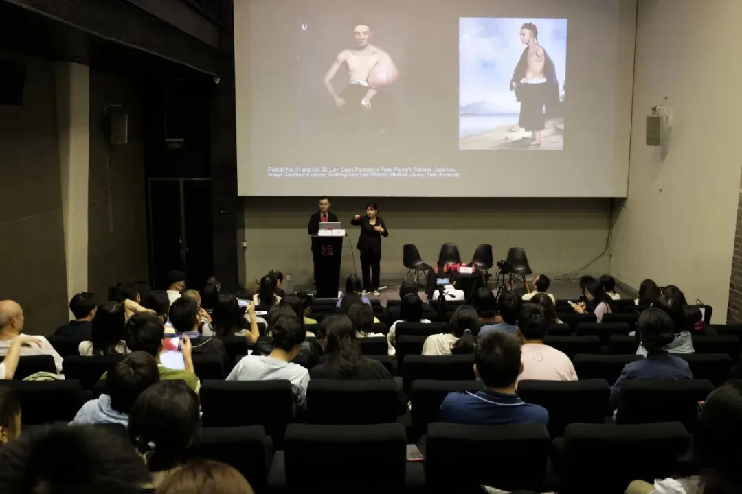 从观众席后方拍摄,大屏幕上展示着此前提到的两幅林官医学肖像画,王超站在讲台前发言,范娇娇在旁打手语,台下坐满观众。