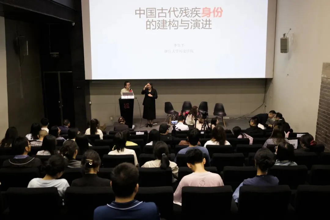 从观众席后方拍摄的报告厅全景,台上李生平站在讲台前发言,手语老师在旁比划,大屏幕显示讲座标题“中国古代残疾身份的建构与演进”,台下坐满观众。