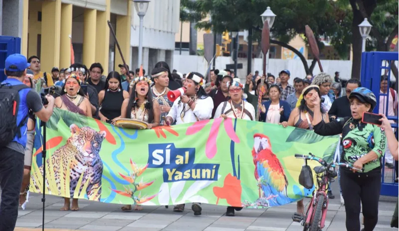 一群头戴传统羽饰的原住民走在街头游行,手持写有“Sí al Yasuní”字样、绘有美洲豹与金刚鹦鹉图案的绿色横幅,两侧民众围观拍照。