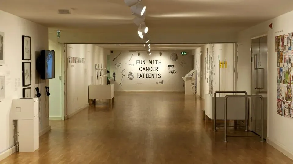 美术馆长廊展览现场,正对面墙上以黑色大字写着“FUN WITH CANCER PATIENTS”,两侧墙面陈列着与治疗相关的手绘图案和展品。