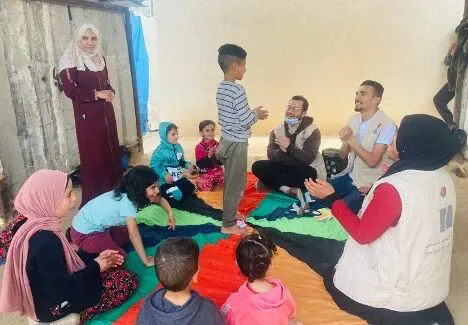 庇护所内几名成人和多名儿童围坐在彩色垫子上做游戏,一名男孩站立在中间,几人戴着口罩或穿着救援机构马甲。