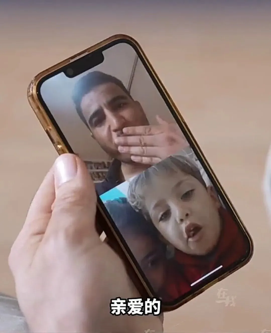 一只手举着手机拍下视频通话画面:一名男子用手捂着嘴,画面另一角是一名孩子,屏幕底部有字幕「亲爱的」。