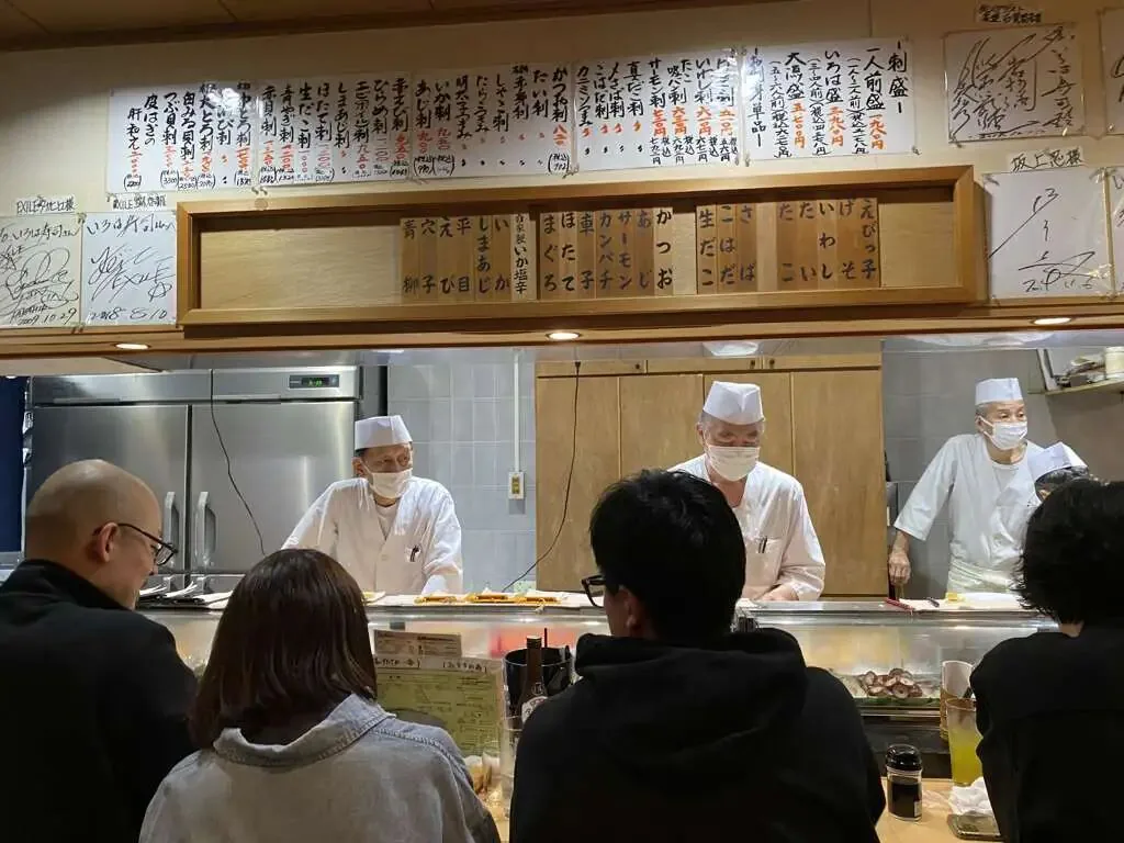 一家寿司店柜台内三位戴口罩的男性厨师在准备食物,头顶挂满手写日文菜单木牌,柜台前坐着几位顾客。
