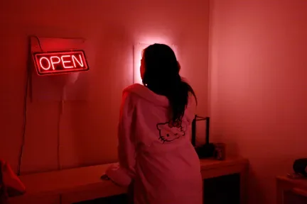红色灯光笼罩的房间里,一人背对镜头站立,身穿印有凯蒂猫轮廓图案的连帽衫,墙上挂着亮着的“OPEN”霓虹灯牌