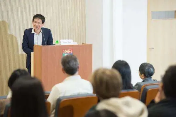 一位身穿深色西装的男性站在讲台后微笑发言,台下听众背对镜头坐在座椅上聆听。