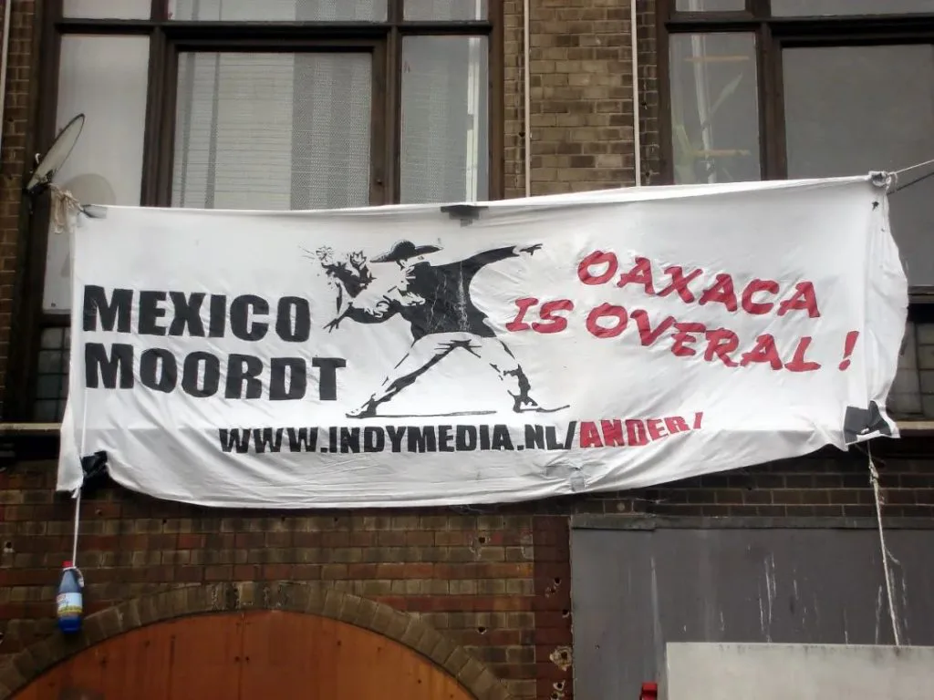 挂在楼房窗前的白色横幅,黑色荷兰语大字「MEXICO MOORDT」(墨西哥在杀戮),红色大字「OAXACA IS OVERAL!」(瓦哈卡无处不在),下方印着网址www.indymedia.nl/ander/,图案是一个持枪者指向持花人的剪影。