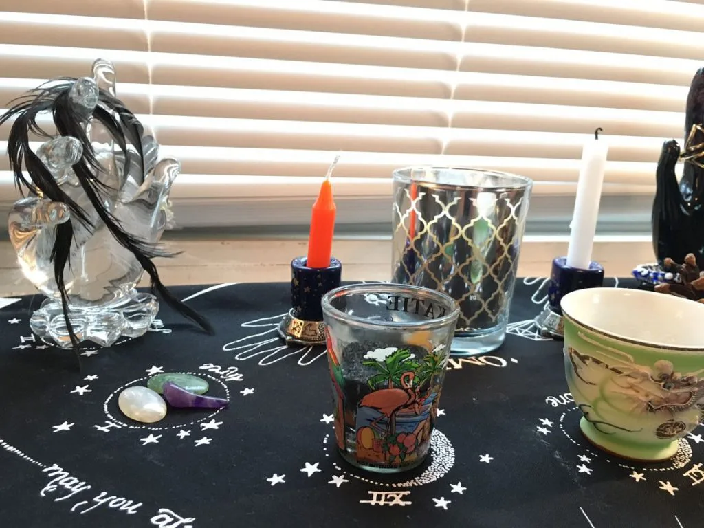 祭坛台面铺着印有星月符号的黑布,摆放一支橙色蜡烛、一尊黑发玻璃女巫摆件、几只印花玻璃杯和一只龙纹瓷碗,桌上还散落着贝壳与松果。