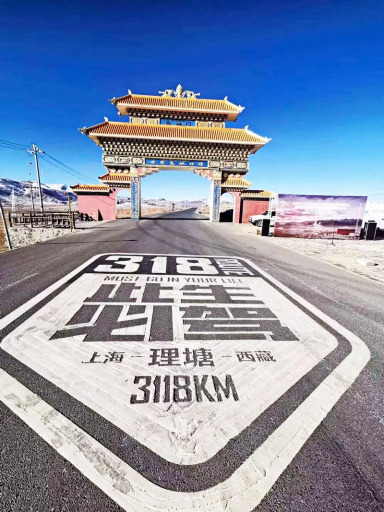 笔直的柏油公路路面喷绘着'此生必驾318 MUST GO IN YOUR LIFE 上海—理塘—西藏 3118KM'字样,远处矗立着一座藏式风格的彩绘牌楼拱门,背景为雪山和蓝天。