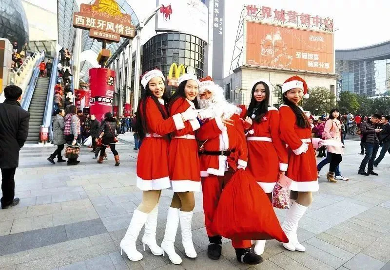 中国商业街上,圣诞老人和几位身穿红色圣诞服饰的女性合影,身后是购物中心大楼和商业街招牌,行人往来。