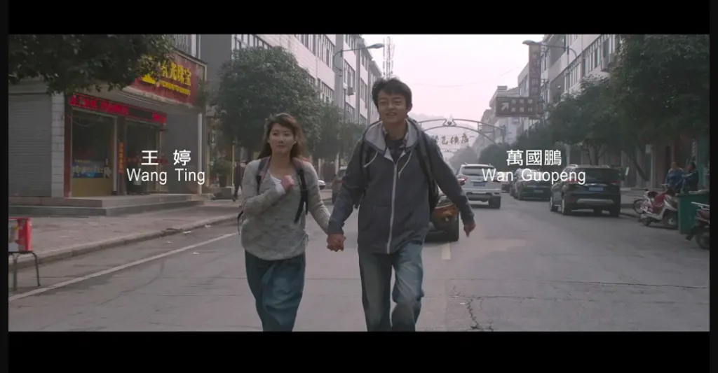 电影片尾画面,字幕标注'王婷 Wang Ting'和'万国鹏 Wan Guopeng',两人牵手走在街道中央,两旁是店铺与楼房。