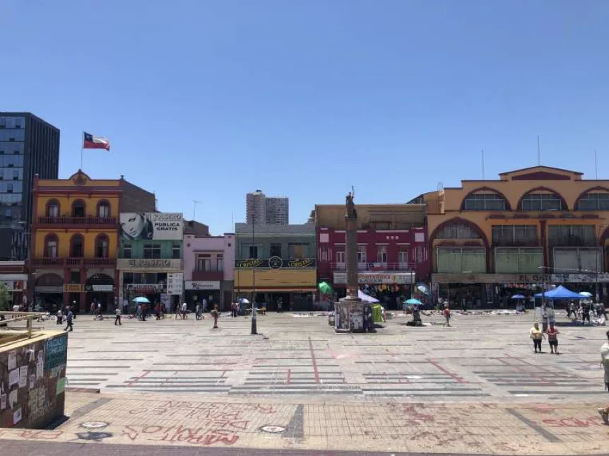 晴天广场全景,地面写满涂鸦,中央一根石柱顶部空置(雕像已不见),四周环绕彩色老式骑楼建筑,零星行人和蓝色帐篷散布广场上。