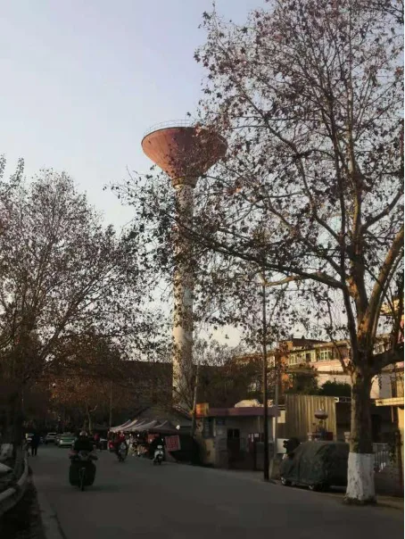 一座高耸的漏斗形水塔矗立在光秃秃的行道树之间,塔身细长、顶部呈伞状展开,背景是老旧居民楼,塔下街道上有行人和摩托车经过。