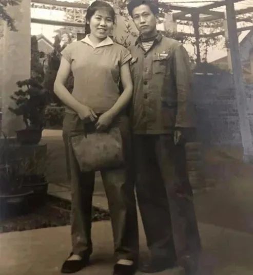 一张黑白老照片,年轻女子留短发、穿短袖衬衫和长裤,手提布包,身旁是穿深色制服式外套的年轻男子,两人站在藤架下,一旁摆着盆景。