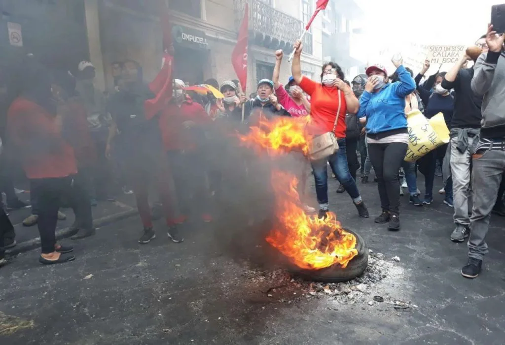 街头一只轮胎燃烧冒出浓烟,周围抗议者举拳呐喊,有人挥舞红旗、举着标语牌,多数人戴口罩。