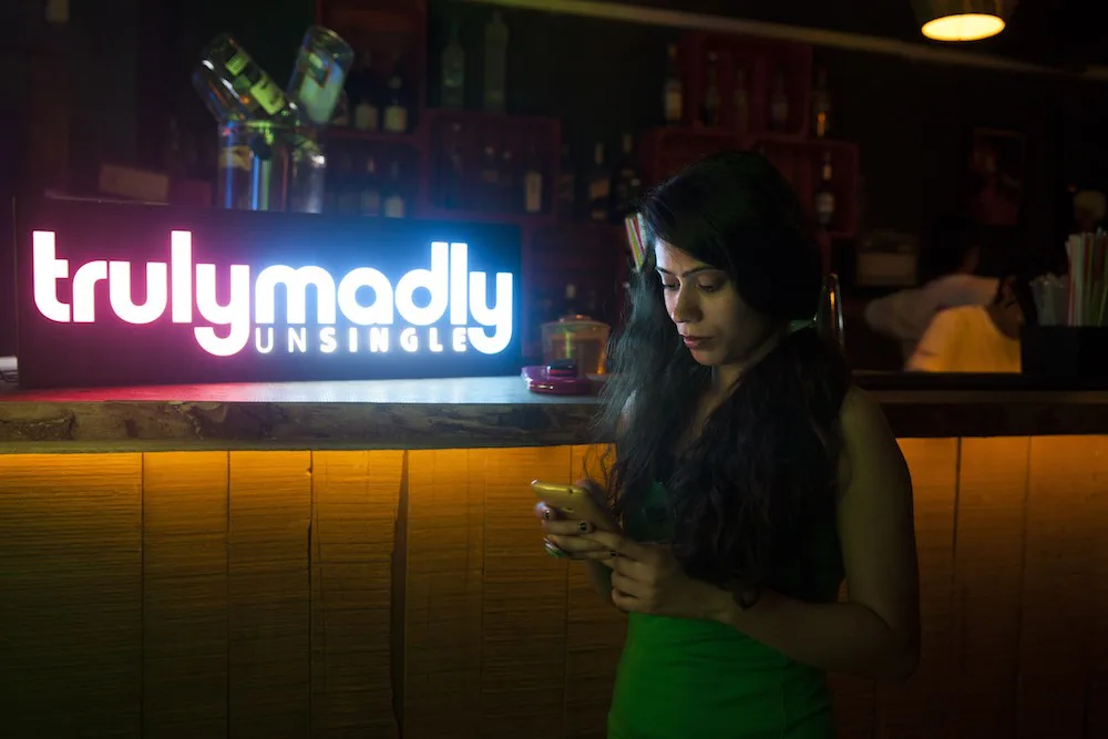 昏暗的酒吧内,一名女子低头专注查看手机,身旁吧台上立着发光的“trulymadly UNSINGLE”约会应用广告牌。