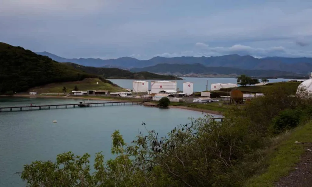 海湾旁的石油仓储罐区,几座白色圆形油罐立在岸边,背后是连绵山脉,海面平静,前景为绿色灌木与山坡。