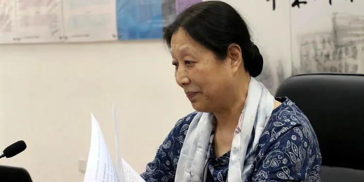 李小江坐在会场上手持稿件发言,面前有麦克风,身穿蓝色印花上衣,肩披围巾。