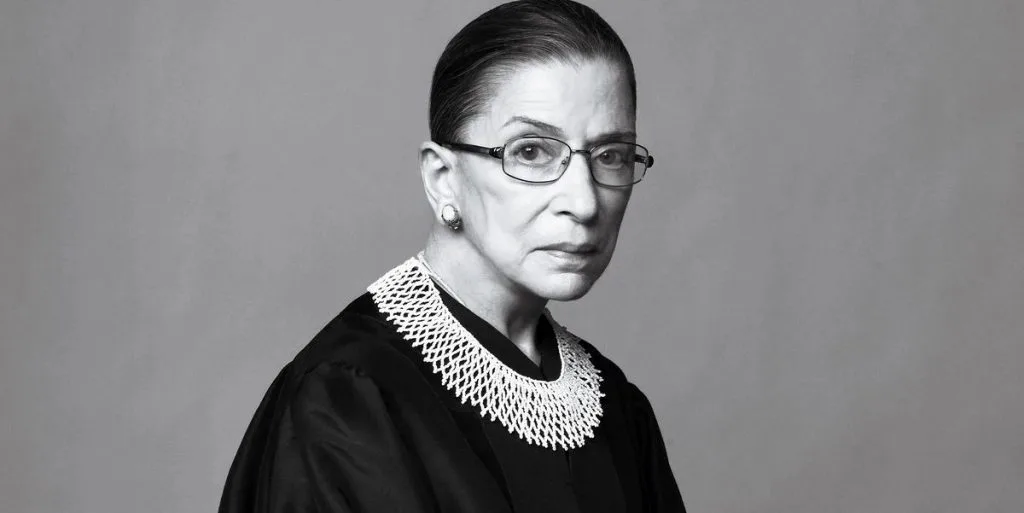 Ruth Bader Ginsburg黑白肖像照,她戴眼镜,身穿黑色法袍,领口佩戴标志性蕾丝领饰。
