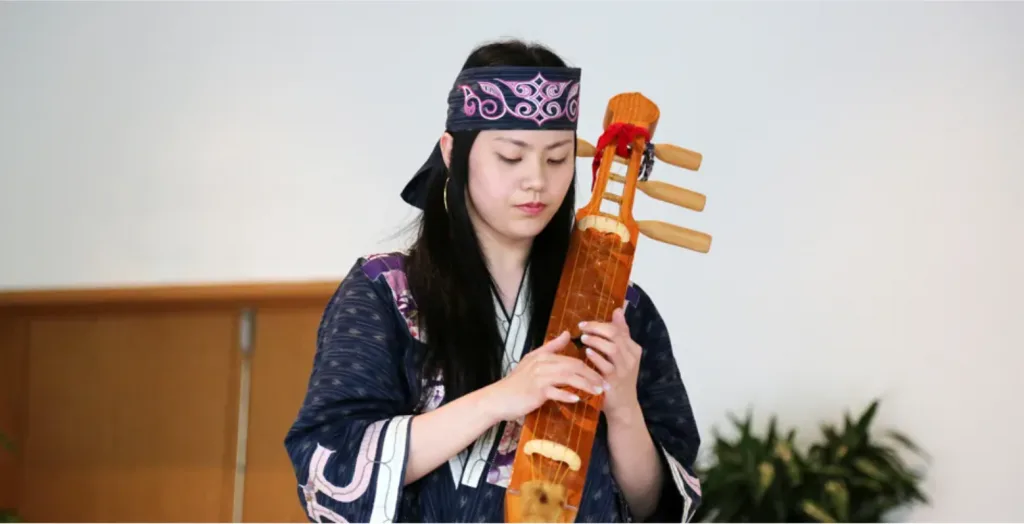 身着深蓝色绣紫白花纹长袍、戴粉色纹样头巾的女性低头专注弹拨竖直握持的木制弦乐器。