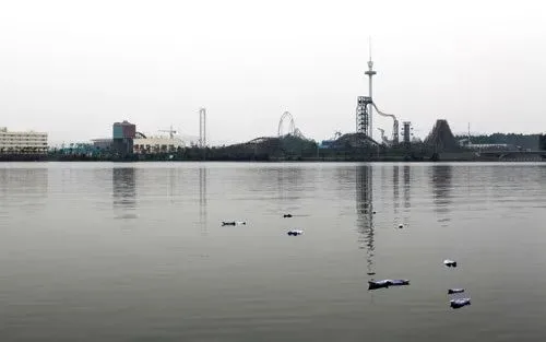 灰蒙蒙的湖面上漂浮着数只白色纸船,远处岸边可见欢乐谷游乐园的过山车轨道与瞭望塔剪影。