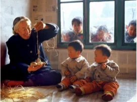 一位老人坐在炕上手持拨楞锤搓麻绳,两个幼儿坐在身旁,窗外几个孩子探头张望。