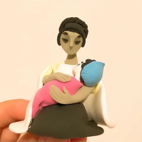 手托着的粘土手办特写:母亲低头凝视怀中裹蓝色毯子的婴儿,神情专注安静。