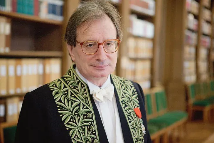 马里翁肖像,戴眼镜,身穿绣有绿色枝叶纹样的学院礼服,站在摆满书籍的图书室内。