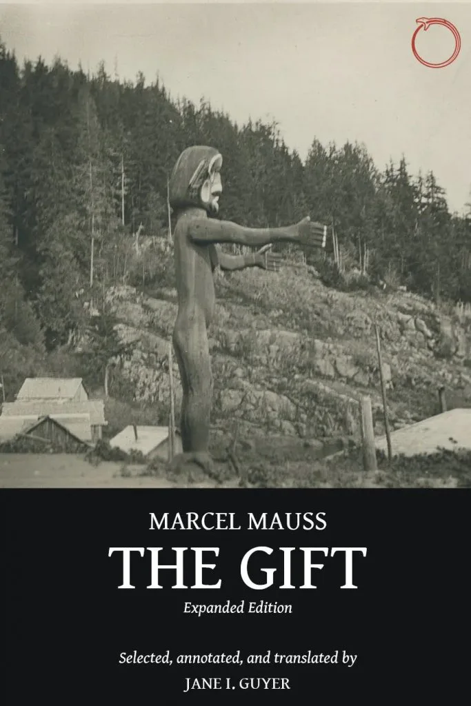 《The Gift》封面,黑白老照片:一座双臂前伸、手持物件的木雕人像立于山坡林间,前方散布几间简易房屋。