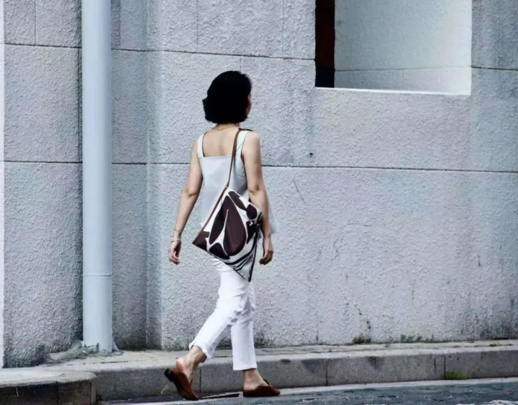 一位身穿白色上衣长裤的女性背对镜头走在灰色墙边,肩上斜挎着印有棕白色曲线图案的包